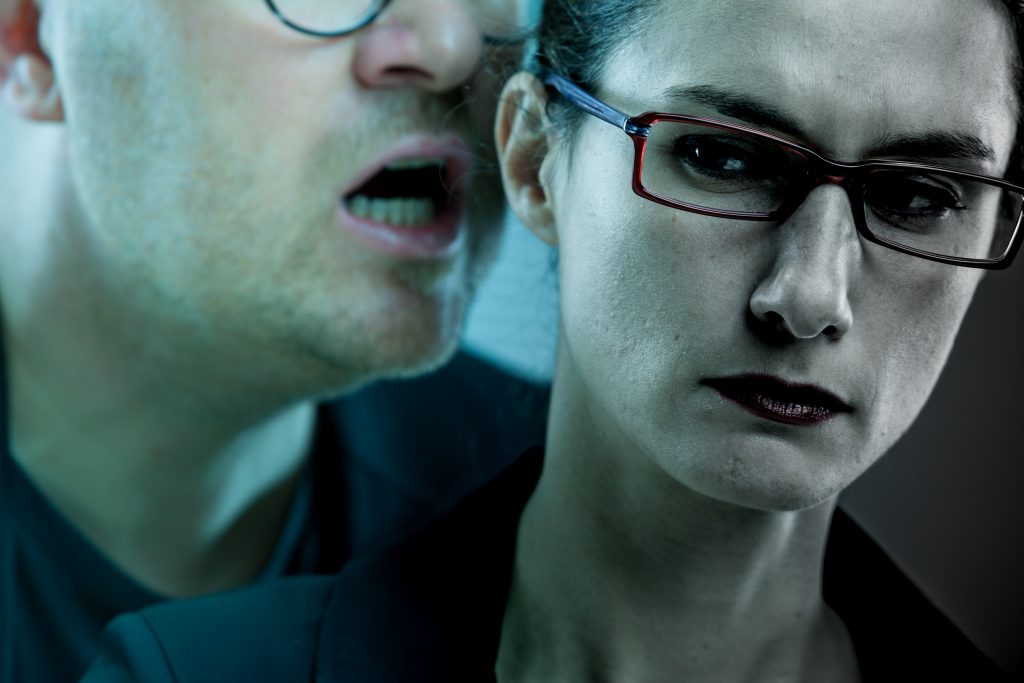 Man with glasses speaking into ear of woman with glasses; woman frowning
